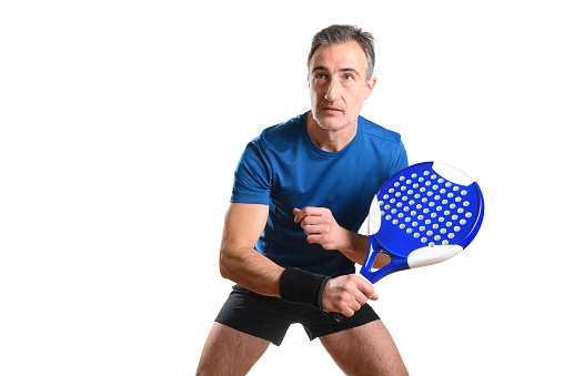 Portrait of man playing padel tennis in position to hit a backhand ball wearing blue and black sports outfit and white isolated background. Front view.