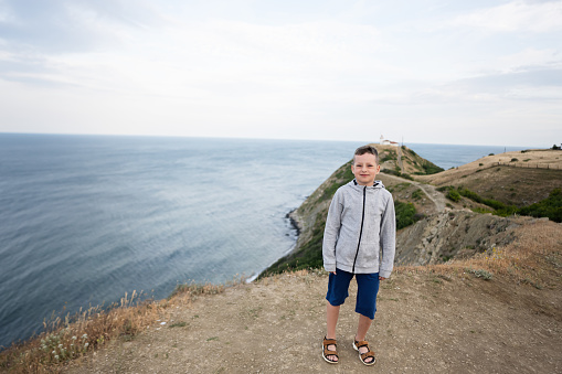 Little boy standing on top of a mountain and looking camera against lighthouse in sea. Cape Emine, Black sea coast, Bulgaria.