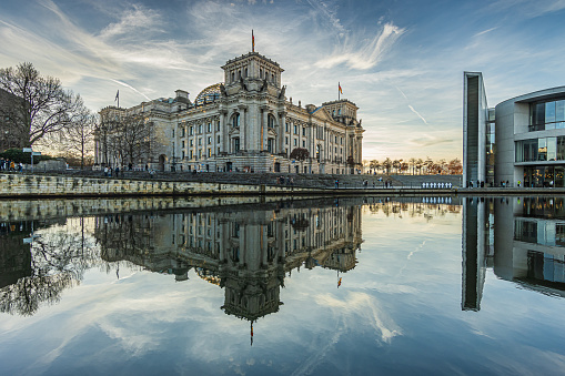historic Reichstag building in Berlin during the day in winter. River Spree with reflection from the building in the government district. Sunshine in the background with cloudy skies.