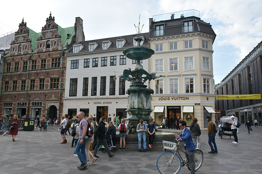 Copenhagen, Denmark - May 15, 2023: Scene Of Copenhagen Shopping District, Fountain, Building Exterior, Advertisement Sign, Retail Store, People Walking Back And Forth, Looking For Items To Buy, Shopping And More During Springtime