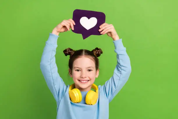 Photo of Photo of positive schoolgirl with buns dressed blue pullover in headphones hold heart icon over head isolated on green color background