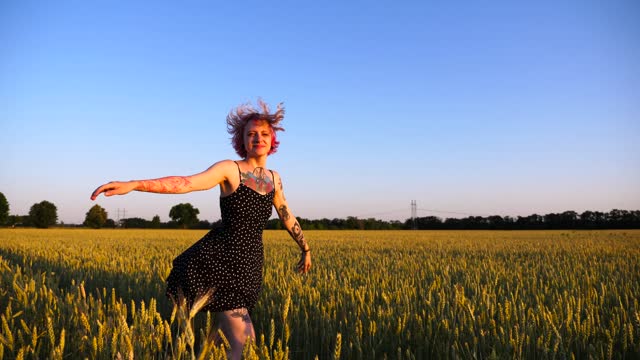 Happy punk woman with tattoos jogging through wheat meadow at sunset. Cheerful hippie girl with pink hair having fun while running among green barley field. Joyful female hipster enjoying summer