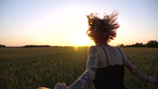 Follow me shot of happy punk girl holds male hand jogging through green wheat field at sunset. Cheerful female hipster pulling her boyfriend among barley meadow. Happy couple having fun at nature. POV