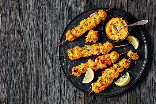 Bang Bang Chicken skewers sprinkled with chives and lemon slices on black plate on dark wooden table, horizontal view from above, flat lay, free space