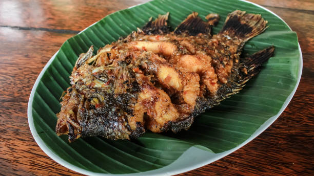 istock Ikan Gurame Bakar or grilled freshwater fish with chilli sauce and vegetable. Serving on white plate on wooden table. Indonesian food and cuisine. Delicious grilled Gouramy. 1635732119