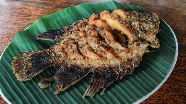 istock Ikan Gurame Bakar or grilled freshwater fish with chilli sauce and vegetable. Serving on white plate on wooden table. Indonesian food and cuisine. Delicious grilled Gouramy. 1635733762