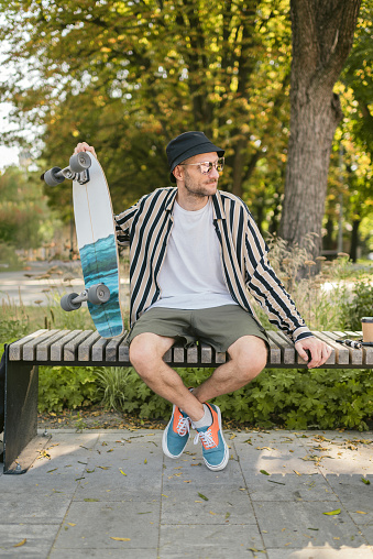 Portrait of stylish adult male who sitting at extreme park near skateboard, summer vibe. resting