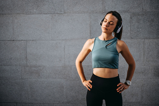Young beautiful athlete woman with wireless headphones warming up for running while standing in front of grey wall outdoors