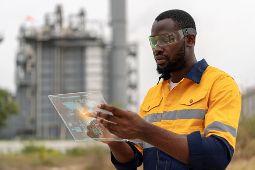 Construction Industry through the lens of an accomplished African American engineer. Dressed in a safety uniform and standing against the backdrop of electrical plant. Expert engineer looking at graphic user interface on the high technology screen and see the new location plan to installation a new power facility.
