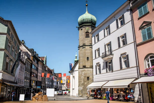 The image showcases a historic tower with a conical, multi-colored tiled roof, surrounded by buildings with red-tiled roofs. The tower, constructed from stone, features small windows and a decorative band near the top. The surrounding buildings have multiple windows with shutters, adding to the architectural charm of the scene.