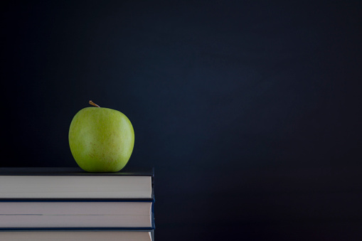 A stack of books with a green apple on the desk on a black background of the blackboard with a place for the inscription.