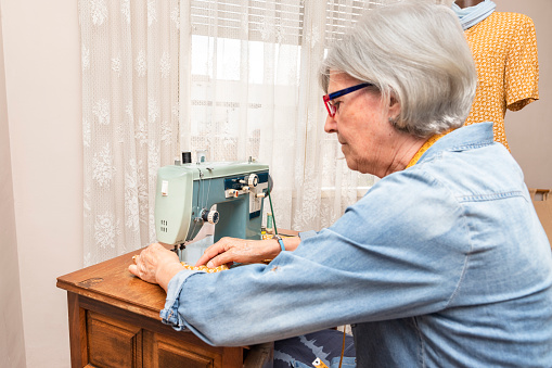 Elderly woman sews in front of an old sewing machine inside a house. horizontal