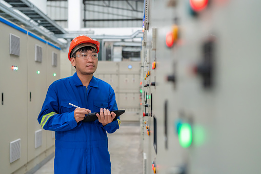 Electric engineer inspects the electric system on a control panel dashboard within a switchgear room. Take note on the clipboard. The careful scrutiny involved in maintaining the integrity of Electrical Systems within the controlled environment of the factory.