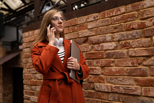 a young female student goes to study in an autumn coat with a laptop and a phone against the backdrop of an urban brick wall.