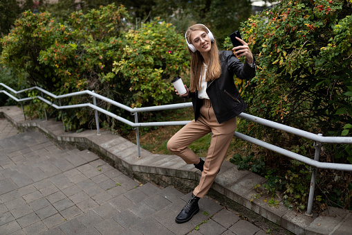 young woman in the park listening to music in headphones with a cup of coffee and taking a selfie.