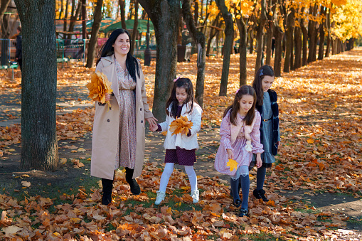mother and daughters are in autumn city park, happy people walking together, family with children, playing with yellow leaves, beautiful nature, bright sunny day