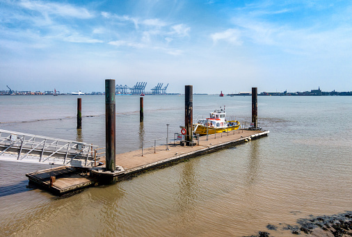 The Harwich Harbour Ferry, with passengers and crew aboard, moored at a pontoon jetty at Shotley Gate in Suffolk, Eastern England. In the distance and to the left are some of the cranes of Port of Felixstowe, Suffolk, and to the right is Harwich in Essex.