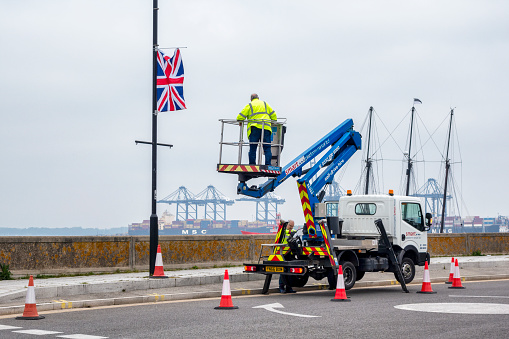 Two workmen, one in a cherry-picker hoist, responsible for taking down celebratory Union Jack flags after the Coronation of King Charles and Queen Camilla. Harwich, Essex, Eastern England, on a cloudy day in May.