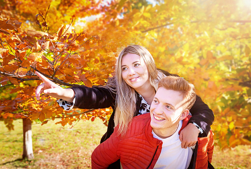 Happy autumn (fall) couple. Boyfriend carrying girlfriend on piggyback. Girl showing with outstretched arm to the side. Young woman, man having fun outdoors. Yellow leaves. Healthy lifestyle.