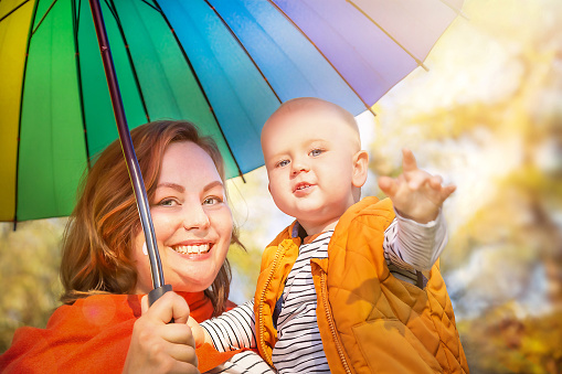Happy emotional baby boy (child, kid) and young mother smiling with colorful umbrella on autumn yellow sunny background.