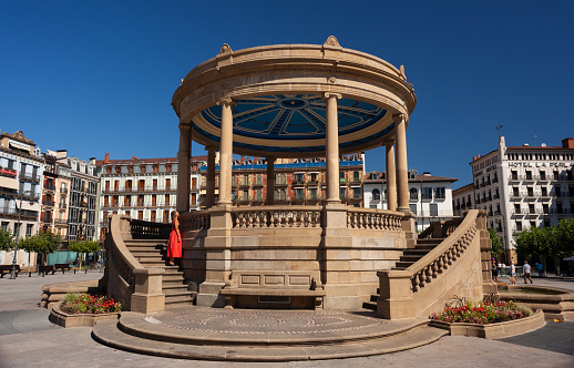 Pamplona, Spain - August 2, 2022: Woman wearing a red dress next to the Pavilion monument at castle Square in the old town of Pamplona Spain.