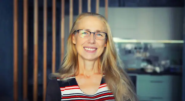 Portrait of happy smiling middle-aged woman in eyeglasses looks at the camera. Senior blonde woman in casual at home.