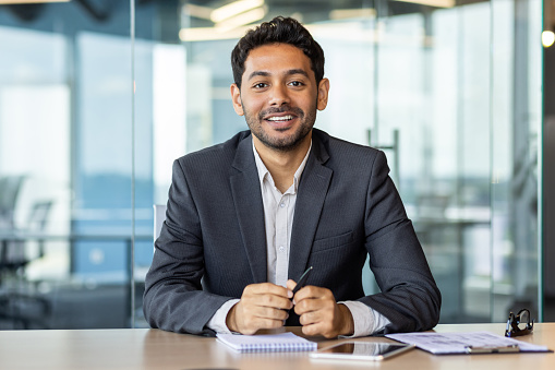 Portrait of young hispanic businessman inside office, boss in business suit smiling and looking at camera, experienced satisfied man at workplace at desk