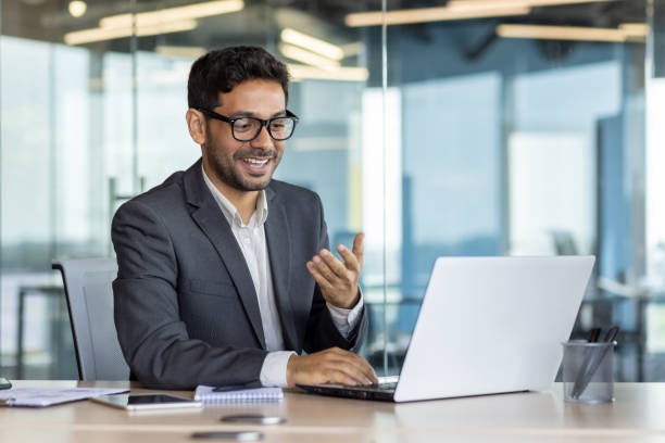 Young successful arab businessman inside office at workplace talking remotely with colleagues and partners, man using laptop for video call, satisfied boss smiling and gesturing with hands joyfully