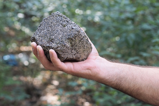 a man's hand holds a large gray piece of stone on a summer street against the backdrop of green vegetation
