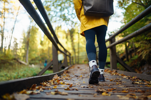 Women's feet in boots go along a wooden walking path in the autumn forest. Vacation travel concept, hiking trail