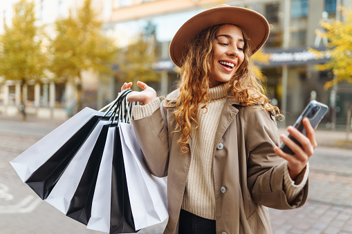 Smiling woman in a coat and hat with bags for shopping, gifts, walking through the sunny streets of the city. Trading woman.