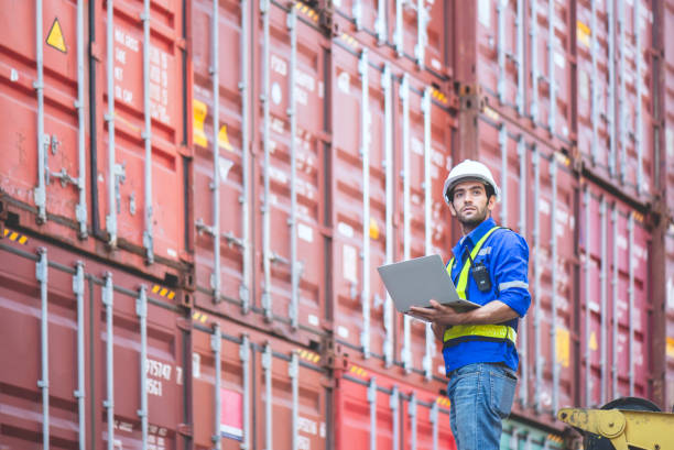 istock Engineer or cargo worker wears PPE standing in front of cargo stacked container yard and holding laptop and looking at the sky. concept logistic delivery management for industrial. 1636175119