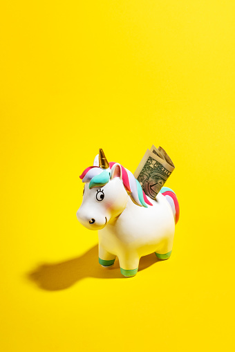 Unicorn shaped coin bank with US dollar banknote. Investment and savings concept. Studio shot on yellow background.