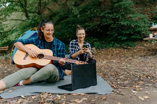 Two women are sitting outdoors in front of their tent, with one of them strumming a guitar and filling the air with delightful melodies