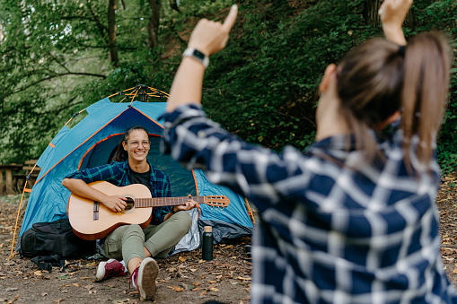 Seated outdoors by their tent, two women enjoy the cozy atmosphere as one of them plays a guitar, infusing the scene with heartwarming melodies