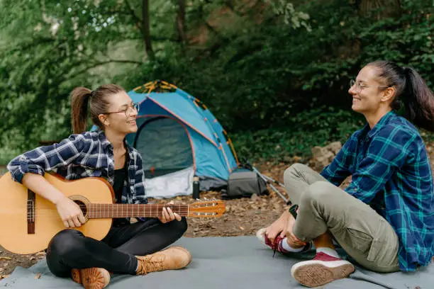 Enjoying the outdoors, two friends sit near their tent, with one of them playing a guitar and filling the surroundings with delightful acoustic tunes