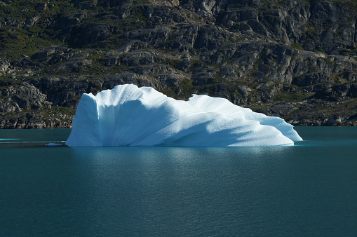 An artwork of a white floating sunlit iceberg with the dark mountains of Greenland in the background in summer
