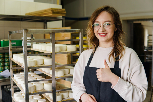 Young smiling woman standing at shelf with cheese heads and showing thumb up.