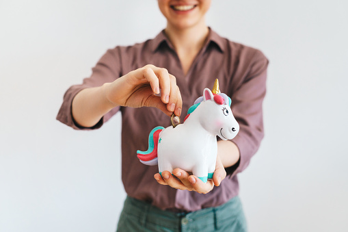 Happy woman putting a coin in a unicorn shaped piggy bank. Start up investment and savings concept.