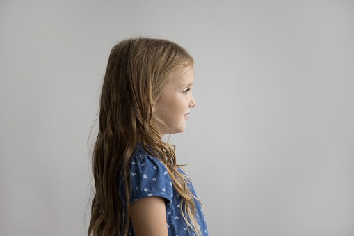 Positive beautiful little kid girl standing at white wall background, looking away, smiling, posing for shooting. Preschool pretty model child with long fair hair side view, profile portrait