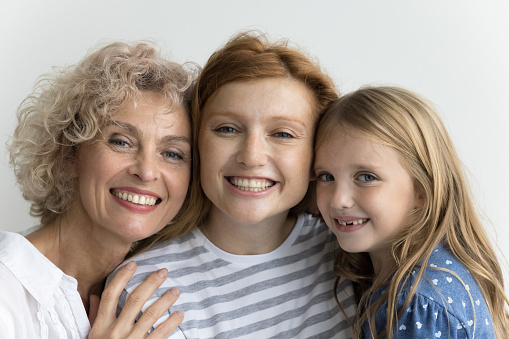 Cheerful cute little kid girl, pretty mom and grandma standing close, hugging with head touches, looking at camera with toothy smiles, promoting family dental care, posing at white wall