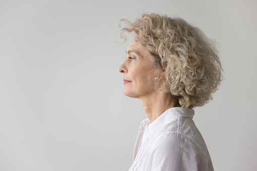 Thoughtful serious pretty blonde mature woman standing at white studio background, looking away in deep thoughts. Beautiful senior model wir curly fair hair side portrait