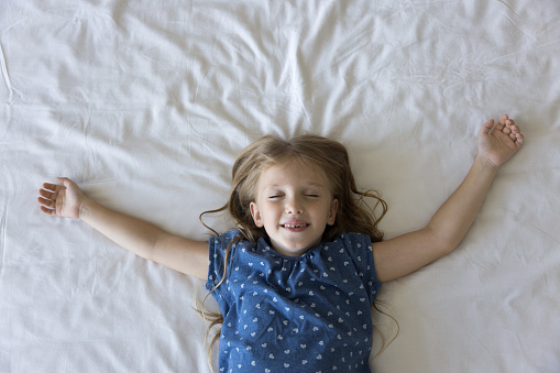 Happy sleepy preschool child girl lying on white sheet on bed with closed eyes, open arms, toothy smile, enjoying relaxation, leisure, comfort on soft orthopedic mattress. Top view