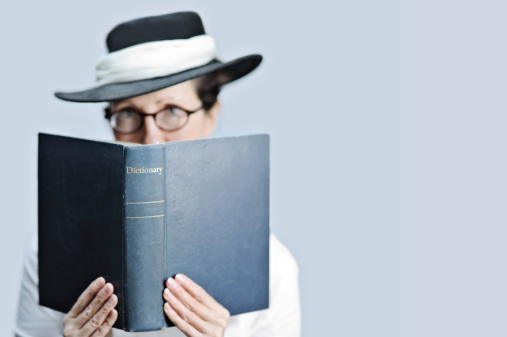 ** Very shallow DOF - focus on word 'dictionary'.** Prim lady librarian/teacher/editor/crossword fanatic/scrabble player/bookworm wearing glasses and Edwardian style hat, holding dictionary and looking at viewer.