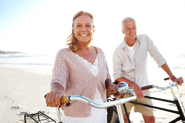 couple souriant tout en prenant un court trajet en vélo sur la plage - senior couple cycling beach bicycle photos et images de collection