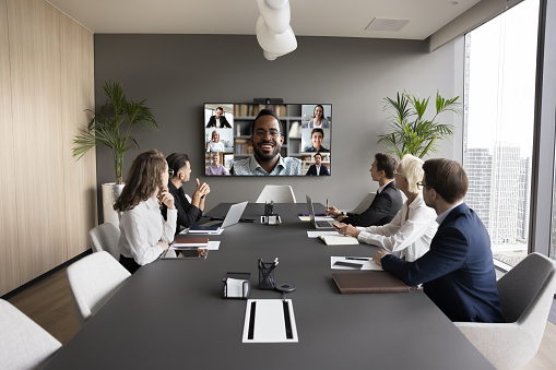 Young African American business leader holding video conference, talking to team online and offline. Workgroup sitting at meeting table, looking at large screen with headshots