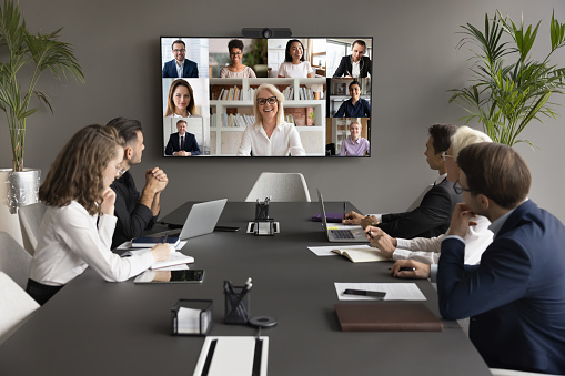 Cheerful mature business mentor woman training diverse office staff on video conference chat, speaking online to diverse freelancers on wall display and office workers at meeting table