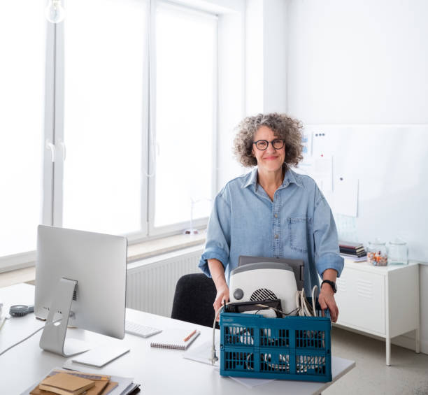 Woman collecting electronic waste in the office Mature woman wearing denim shirt standing in the office and holding basket with e-waste, smiling at camera. laptop in a trash bin stock pictures, royalty-free photos & images
