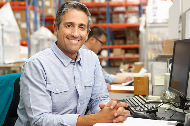 Businessman Working At Desk In Warehouse Businessman Working At Desk In Warehouse Smiling To Camera dispatcher stock pictures, royalty-free photos & images