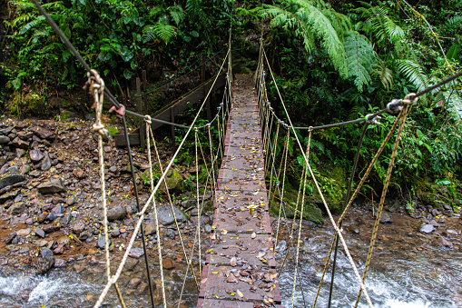 Wide angled, high point photo of a hanging rope bridge over a shallow forest river. Trees with lush foliage are visible in the background, with the bridge in the center and the rocky shore below. Photo taken in El Tigre, Monteverde Cloud Forest in Costa Rica.